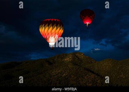 Colourful glowing hot air balloons floating in a night sky over forested mountain peaks