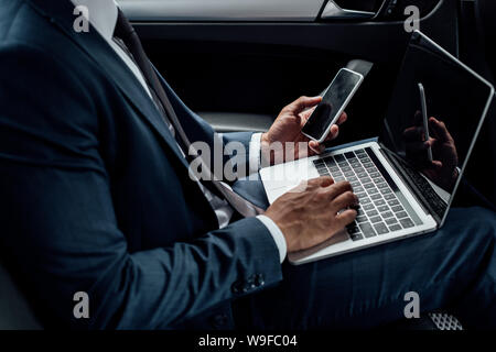 cropped view of african american businessman using laptop and smartphone in car Stock Photo