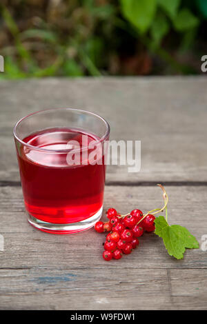 branch of red currant berries and a glass Cup with delicious berry juice on the edge of an old wooden table in the morning Summer garden. Stock Photo