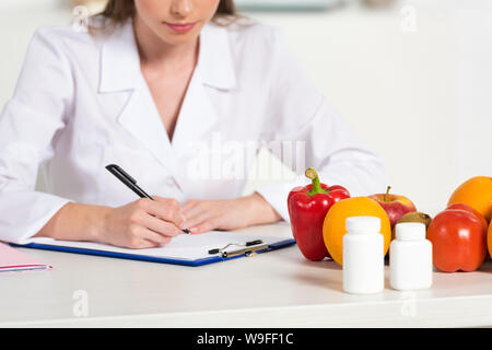 cropped view dietitian in white coat writing in clipboard at workplace with pills, fruits and vegetables on table Stock Photo