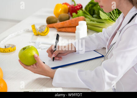 cropped view of dietitian in white coat holding green apple and writing in clipboard Stock Photo