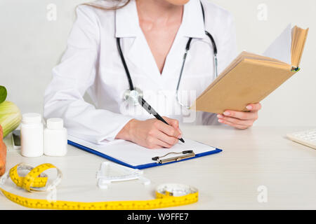 cropped view of dietitian in white coat reading book and writing in clipboard at workplace Stock Photo