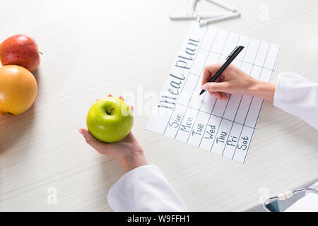 cropped view of dietitian holding apple and writing in meal plan at workplace Stock Photo