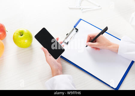 cropped view of dietitian holding smartphone with blank screen and writing in clipboard Stock Photo