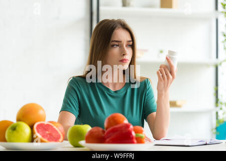 sad woman sitting at table with fruits and vegetables and holding pills Stock Photo