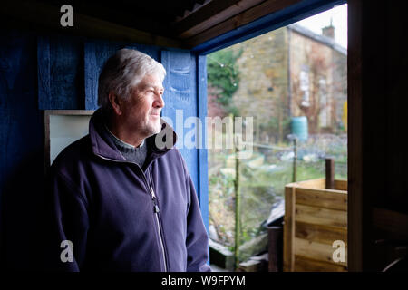 A man in his late sixties / early seventies looks out from his blue garden shed to his home garden and compost heap on a rainy winter day in Northumbe Stock Photo