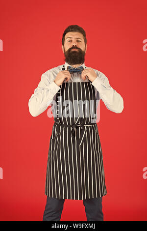 His style works really well to compliment beard. Bearded man fixing bow tie in bib apron. Elegant hipster with bearded face. Bearded bartender or cook in work uniform. Long bearded waiter or servant. Stock Photo