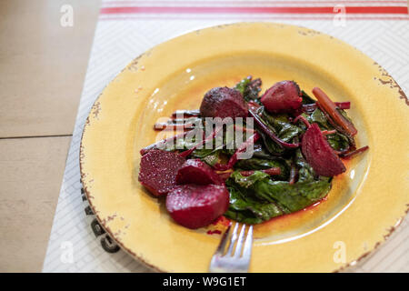 Hot fresh cooked beets or beetroot with leafy tops, seasoned with olive oil and garlic on a yellow plate. Stock Photo
