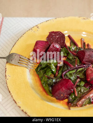 Hot fresh cooked beets or beetroot with leafy tops, seasoned with olive oil and garlic on a yellow plate. Stock Photo