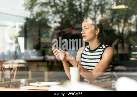 Thoughtful caucasian woman holding mobile phone while looking through the coffee shop window during coffee break. Stock Photo