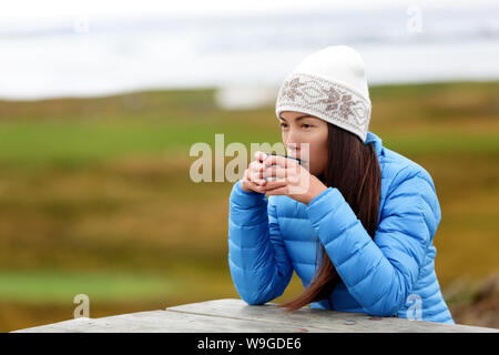Woman in outdoors drinking coffee from thermos cup sitting outside wearing warm down jacket and knit hat. Pretty young mixed race Asian Chinese Caucasian woman living active lifestyle. Stock Photo
