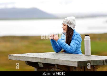 Woman in outdoors drinking coffee from thermos bottle flask sitting outside wearing warm down jacket and knit hat. Active young mixed race Asian Chinese Caucasian woman enjoying traveling on Iceland. Stock Photo