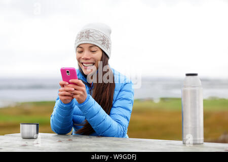 Woman outdoors using smartphone app on smart phone drinking coffee from thermos cup sitting outside wearing warm down jacket. Pretty young mixed race Asian Chinese Caucasian woman in active lifestyle. Stock Photo
