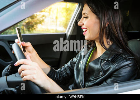 Happy smiling woman holding her smart phone sitting inside a car. Female driver laughing reading message and chatting in mobile. Girl looks at her Stock Photo