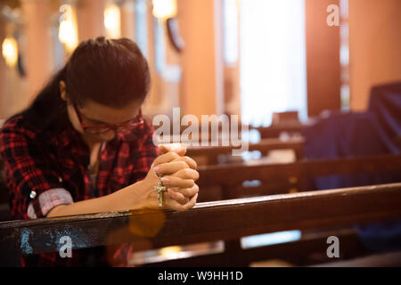 Young woman sits on a bench in the church and prays to God. Hands folded in prayer concept for faith. Stock Photo