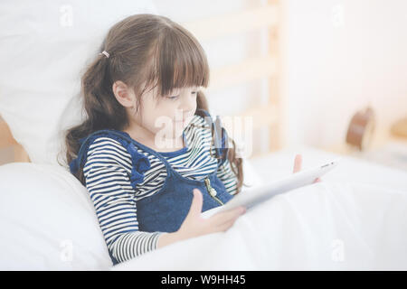 Cute asian little girl enjoy watching cartoon on smart tablet while sitting on bed in kid's bedroom at home. Stock Photo
