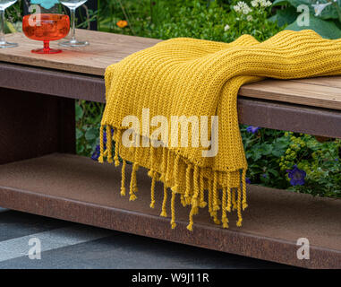 Outdoor wood and metal garden bench on grey stone patio area with mustard colour fabric throw and glasses Stock Photo