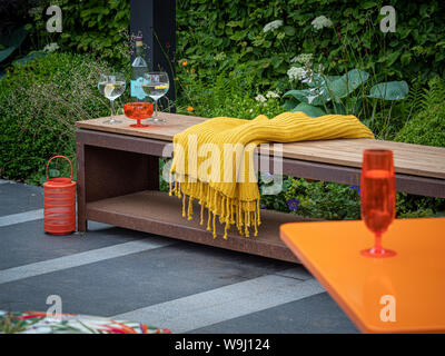 Outdoor wood and metal garden bench on grey stone patio area with mustard colour fabric throw and glasses Stock Photo