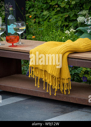 Outdoor wood and metal garden bench on grey stone patio area with mustard colour fabric throw and glasses Stock Photo