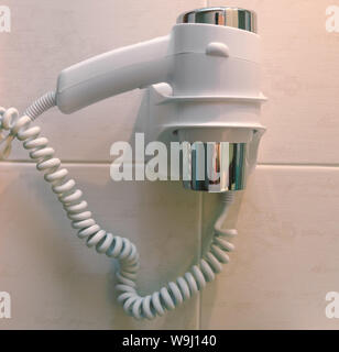 A small compact hair dryer in a holder attached to the bathroom wall. Stock Photo