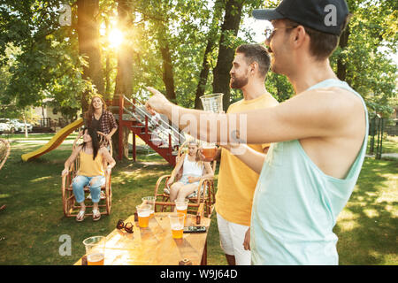 Group of happy friends having beer and barbecue party at sunny day. Resting together outdoor in a forest glade or backyard. Celebrating and relaxing, laughting. Summer lifestyle, friendship concept. Stock Photo