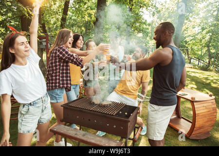 Group of happy friends having beer and barbecue party at sunny day. Resting together outdoor in a forest glade or backyard. Celebrating and relaxing, laughting. Summer lifestyle, friendship concept. Stock Photo
