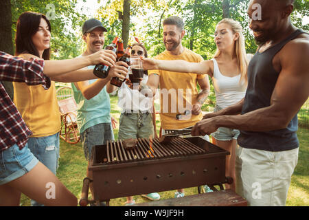 Group of happy friends having beer and barbecue party at sunny day. Resting together outdoor in a forest glade or backyard. Celebrating and relaxing, laughting. Summer lifestyle, friendship concept. Stock Photo