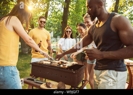 Group of happy friends having beer and barbecue party at sunny day. Resting together outdoor in a forest glade or backyard. Celebrating and relaxing, laughting. Summer lifestyle, friendship concept. Stock Photo