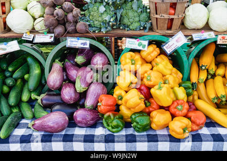 colorful produce on a table covered with a blue and white checkered tablecloth including eggplant, yellow, red, orange, and green bell peppers as well Stock Photo