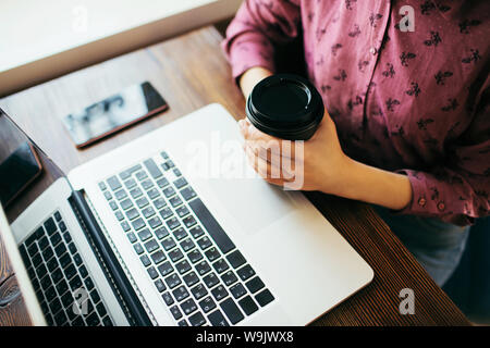 Overview of a young woman working in a cafe in front of laptop, holding a cup of coffee. Smartphone beside. Stock Photo