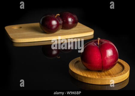 Group of three whole ripe red round plum on bamboo cutting board on round bamboo coaster isolated on black glass Stock Photo