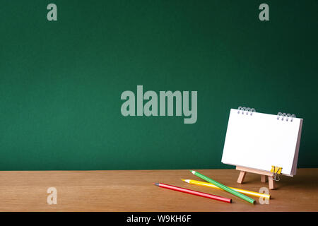 Back to school concept, open notebook on miniature easel and several colored pencils on wooden surface on the backdrop of a clean green chalk board, s Stock Photo