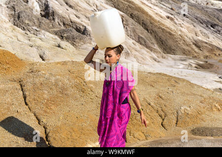 girl of oriental appearance in a sari carries on her head a large plastic canister  with water over a desert area Stock Photo