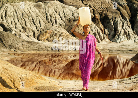 girl of oriental appearance in a sari carries on her head a large plastic canister  with water over a desert area Stock Photo