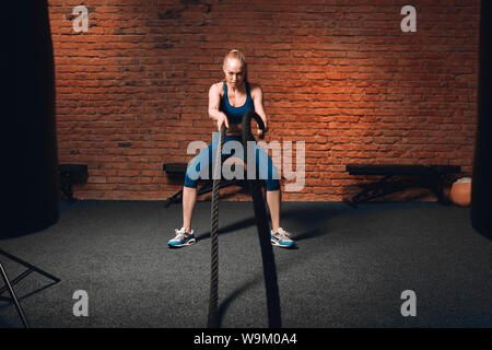 fair-haired beautiful serious athletic woman doing battle rope exercises at gym, full length photo. health and body care. Stock Photo