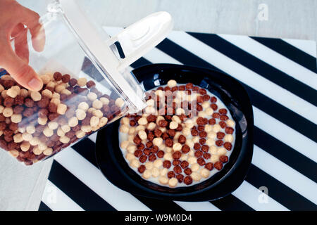 female hand fills from the container chocolate cereal and corn balls in a black plate with milk. Stock Photo