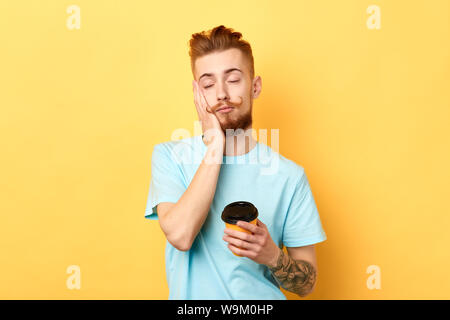 Tired exhausted guy with sleepy expression, covers face with palm, holds disposable paper cup of coffee. isolated yellow background Stock Photo
