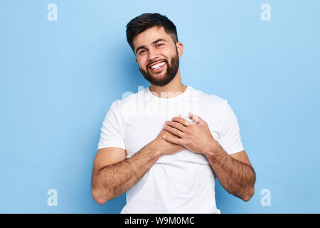 happy overjoyed hipster guy smiles happily, keeps hands on heart, expresses pleasant emotions sympathy and great love.close up portrait, man has falle Stock Photo