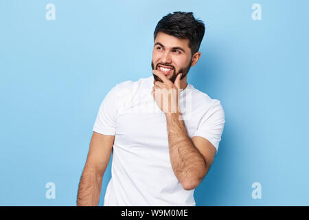 Young handsome guy wearing a white t-shirt, holds his hand at his chin and looks thoughtfully aside isolated over blue background.young man planning h Stock Photo