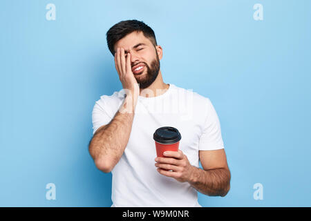 Tired exhausted man covers face with palm, holds disposable paper cup of coffee. man doesn't want to go to work. isolated blue background, tiredness, Stock Photo