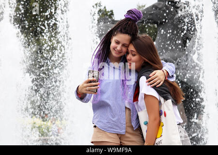 Two hugging girls take a selfie on smartphone on background of splashing fountain Four Seasons on Manezhnaya square. Friendship of diverse ethnicities Stock Photo