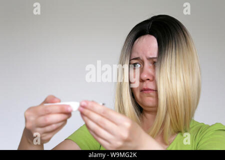 Upset woman looks at digital thermometer in her hands. Measuring body temperature, concept of fever, cold and flu, illness Stock Photo