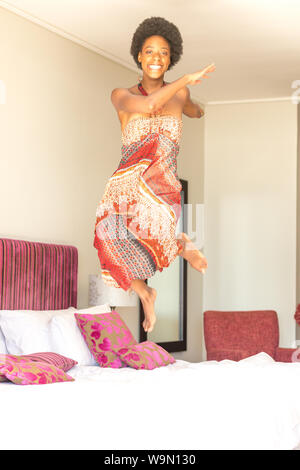 Young African woman jumping on a luxurious bed playfully in a summer dress Stock Photo