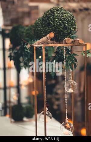 shot of candles inside of round glasses hanged on chains under green plants on a wooden stand Stock Photo