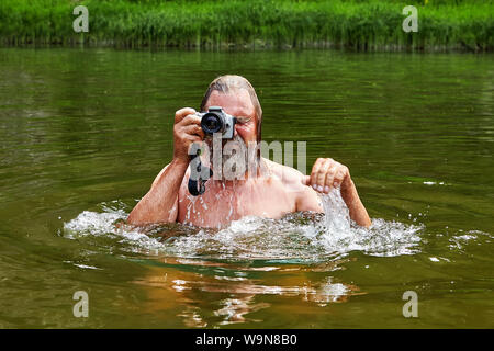Wet mature bearded man is standing in water and taking pictures by digital camera. Stock Photo