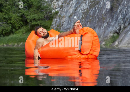 A young businessman, during his vacation, fell asleep in an inflatable lounger and drifting along the river, lowering his hand with the phone into the Stock Photo