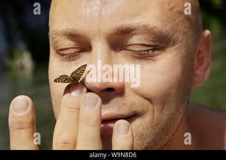 Beautiful butterfly is sitting on the nose of cheerful hairless man, he is touching it by his fingers, close-up. Stock Photo