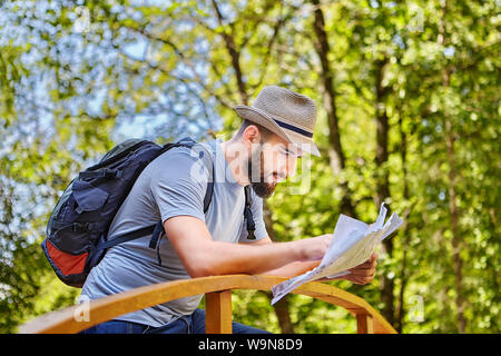 Young pretty caucasian backpacker is checking a map of his route being on the bridge in forest. Stock Photo