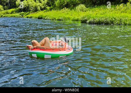 Young attractive caucasian man with hat on his head is drifting down the river in forest on the inflatable ring. Stock Photo