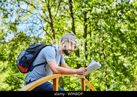 Young pretty caucasian man with hat on his head and backpack is checking map on the bridge in forest. Stock Photo